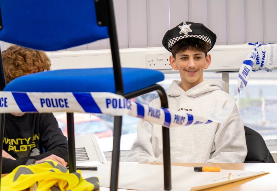Student wearing a police hat in a classroom surrounded by police tape