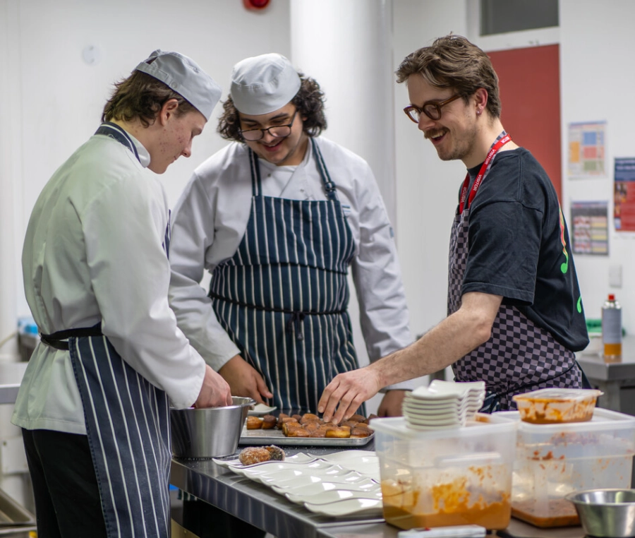 3 males preparing food food in a kitchen smiling and wearing their chefs uniforms