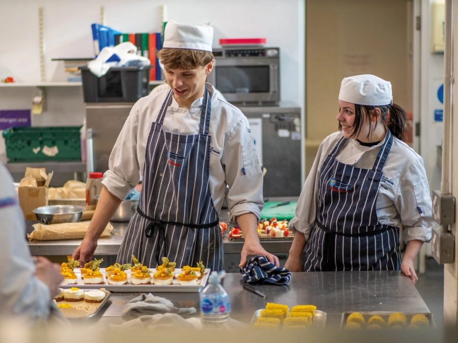 Two students wearing chefs uniform preparing food in kitchen
